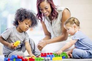 A teacher is sitting on the floor with two preschool children - they are playing with colorful lego blocks.